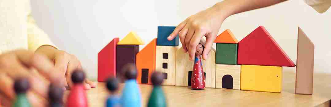 A child's hand placing a small wooden figure among a row of brightly coloured block houses.