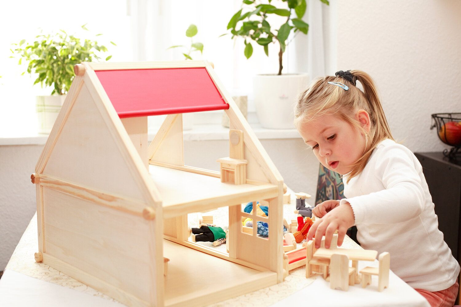 A girl kneeling at an open wooden dolls house with a red roof, arranging tiny furniture inside it.