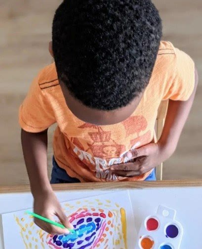 A boy seen from above, painting a bright spiral picture beside a set of watercolours.