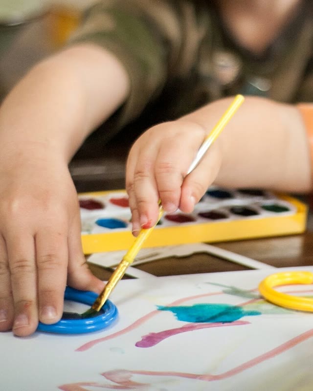 A child's hands steadying a paint pot while painting with a yellow brush.
