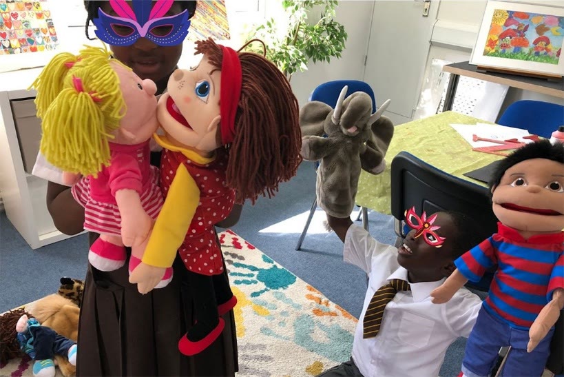 Children in a play room holding soft puppets, two of them wearing bright feathered masks.